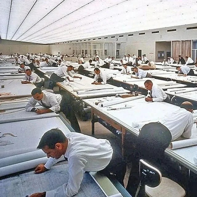 A mid-century drafting hall with rows of drafters at long tables.
